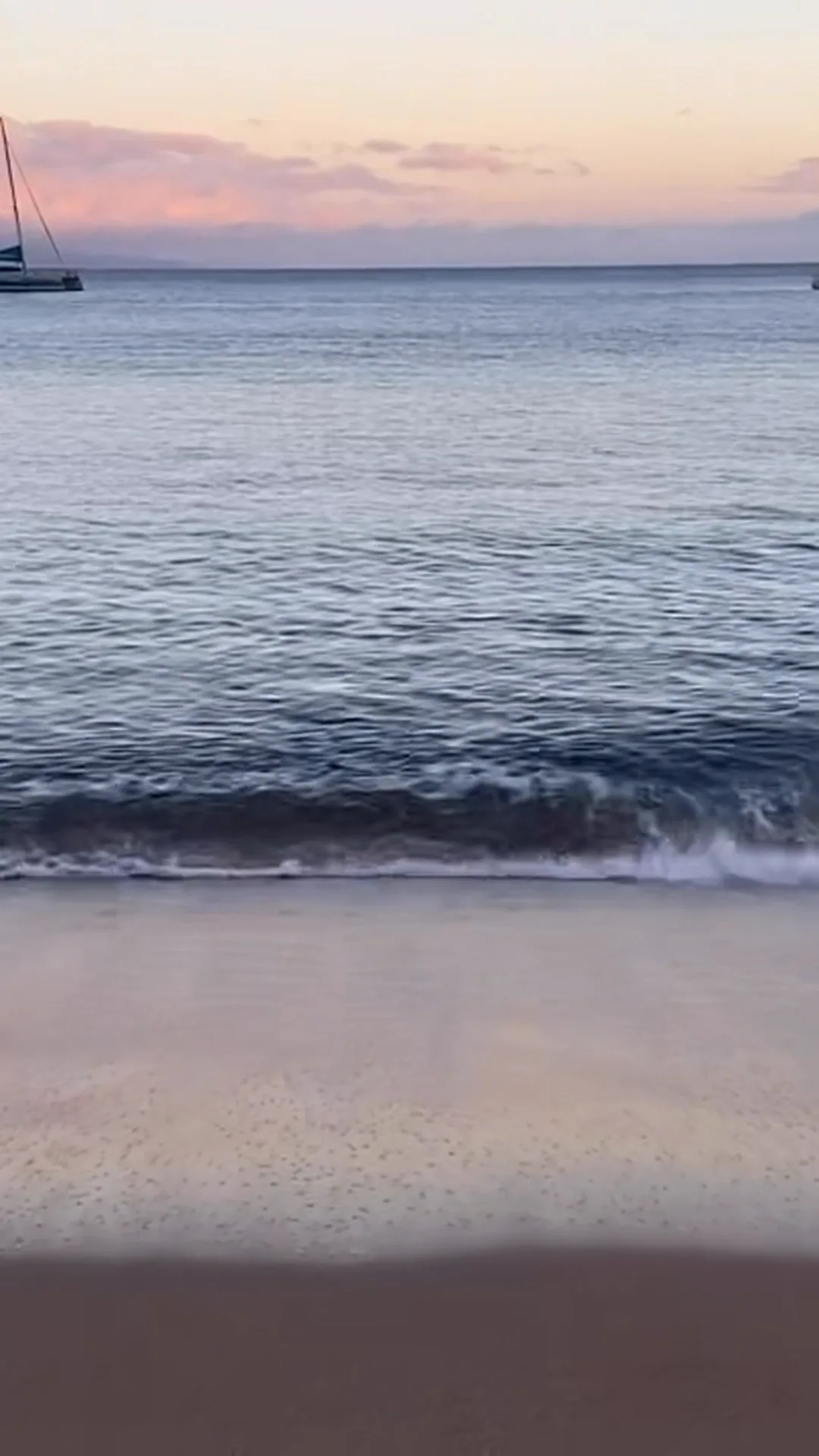 Dawn over a calm sea: a small wave breaking into foam above wet sand that mirrors the pastel sky, a catamaran resting at anchor at the edge of the frame