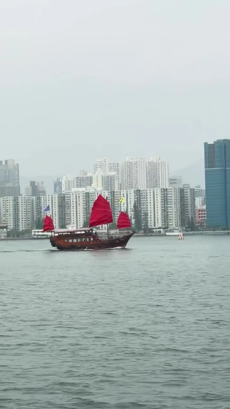 A red-sail junk with residential towers behind it