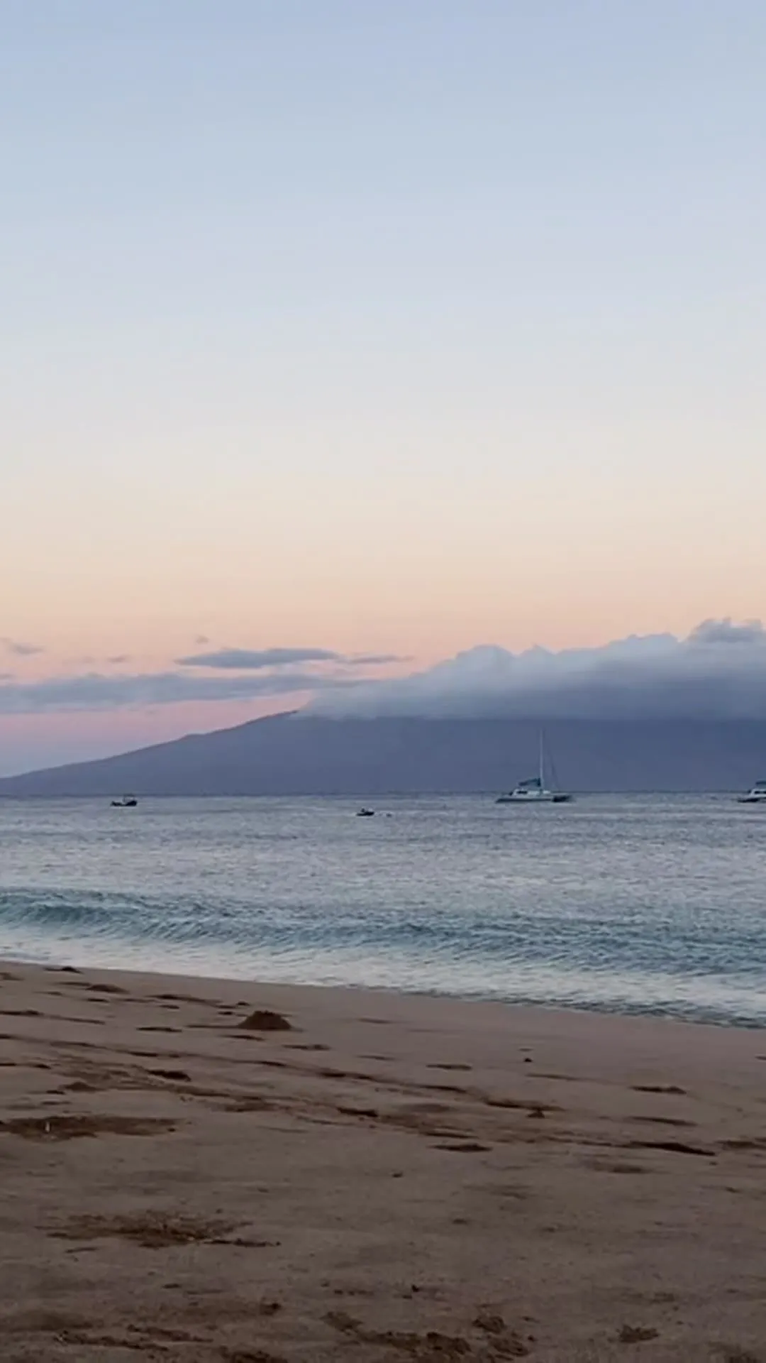 夏威夷海灘外,日出時分海面上的一艘帆船;海峽對岸的島嶼還沉在粉紅色晨空下的暗影裡