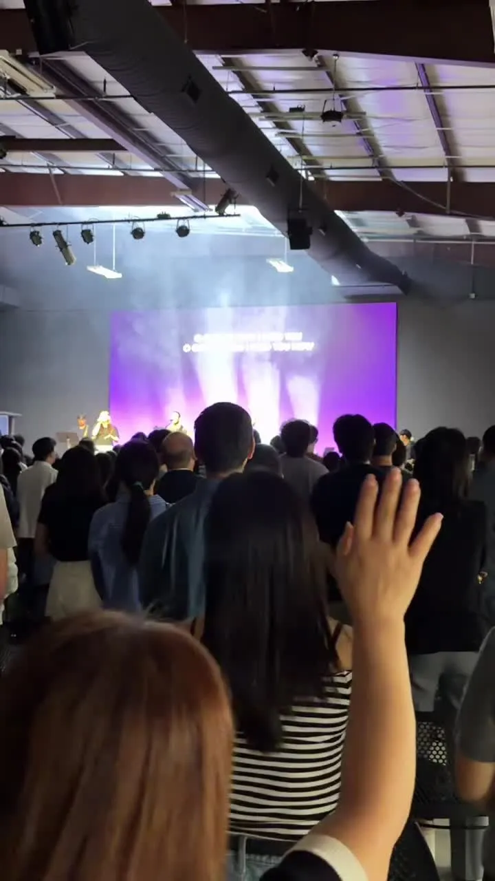 A young woman in the congregation raises her hand in worship, seen from the back row; the screen glows purple with the words of the song