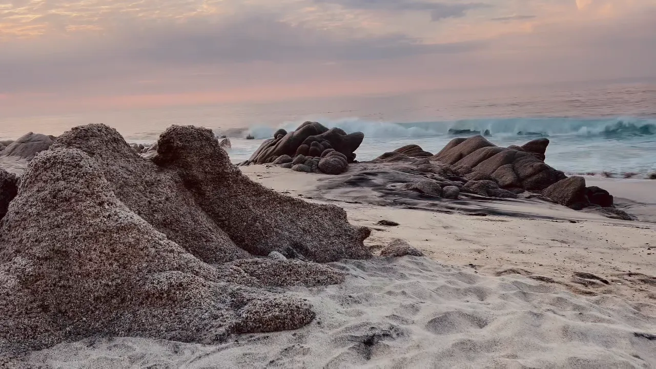 Cabo 海岸上被海浪磨圓的花崗岩巨石,身後一波浪正碎開,頭上是粉紅色的晚空