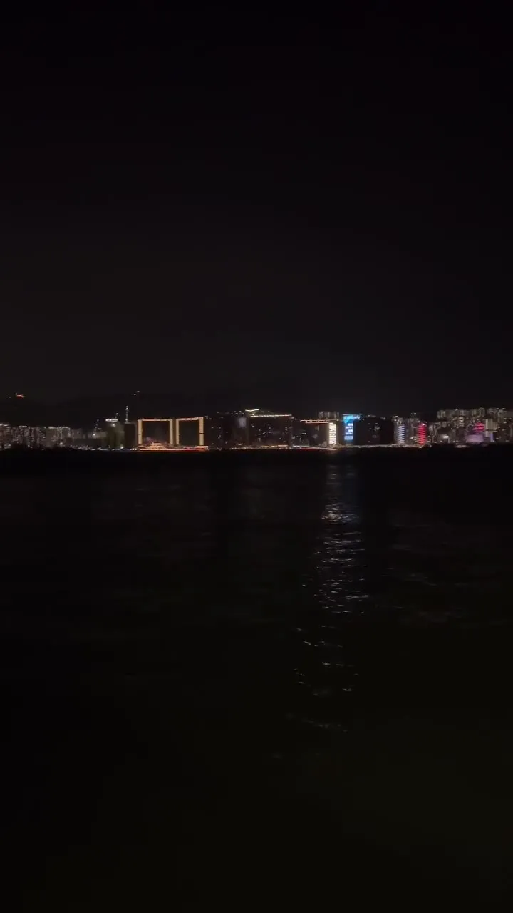 La orilla lejana del puerto Victoria encendida de punta a punta de noche: torres perfiladas y letreros de colores sobre el agua negra