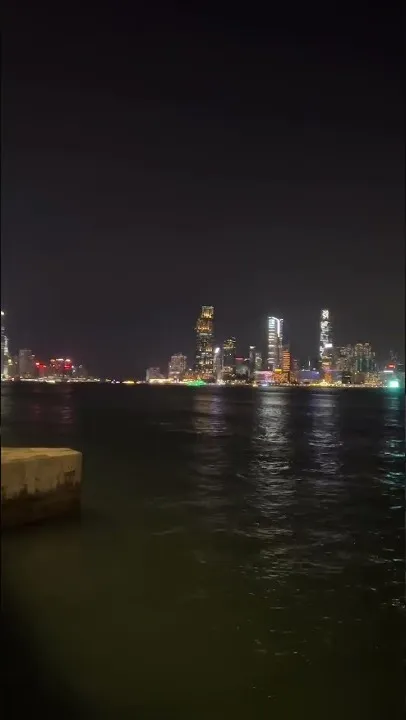 El horizonte de Hong Kong al otro lado del agua de noche: torres encendidas y su luz rompiéndose en la superficie del puerto