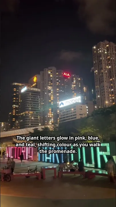 Las letras gigantes de VICTORIA HARBOUR brillando en rosa y verde azulado en el paseo de noche, con gente paseando