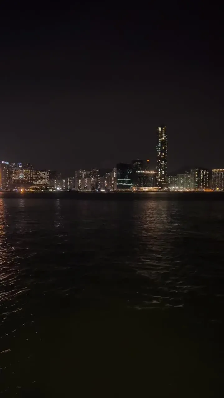 Un muro de torres encendidas al otro lado del puerto Victoria de noche, con una torre esbelta por encima de todas