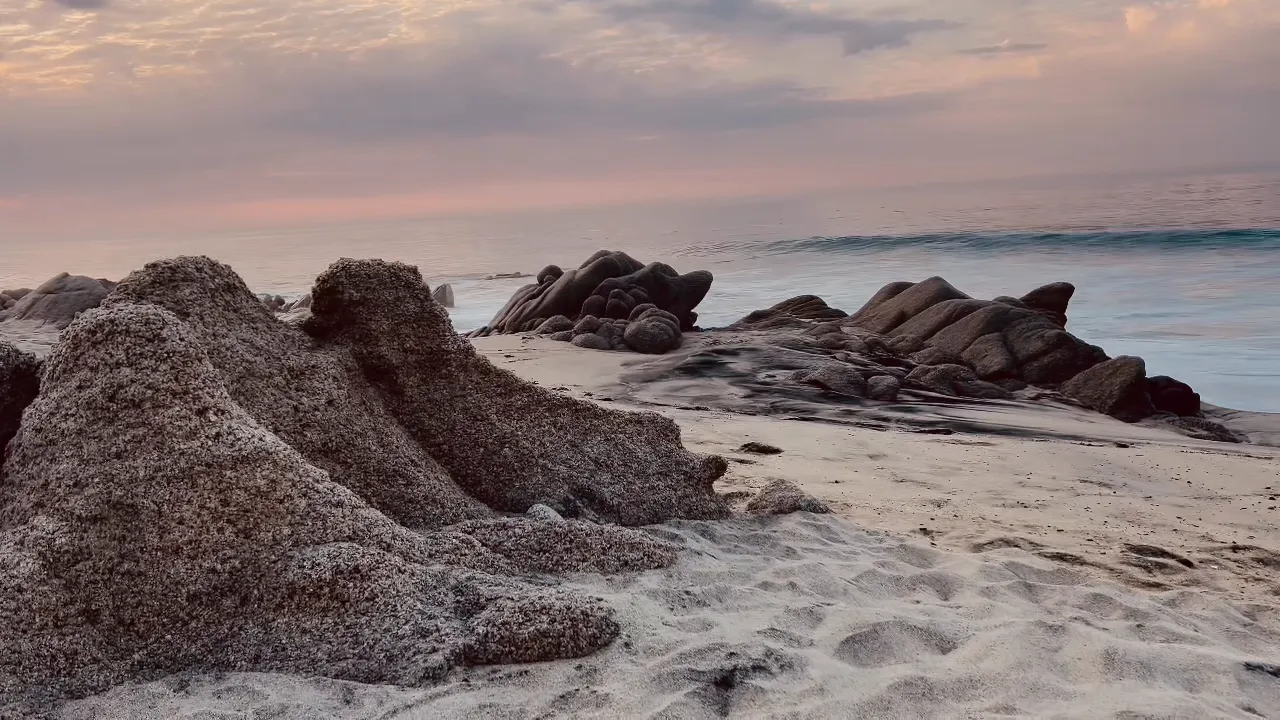 黃昏的 Cabo 海岸,花崗岩礁石立在沙上,身後是粉彩色的天空與平靜的太平洋
